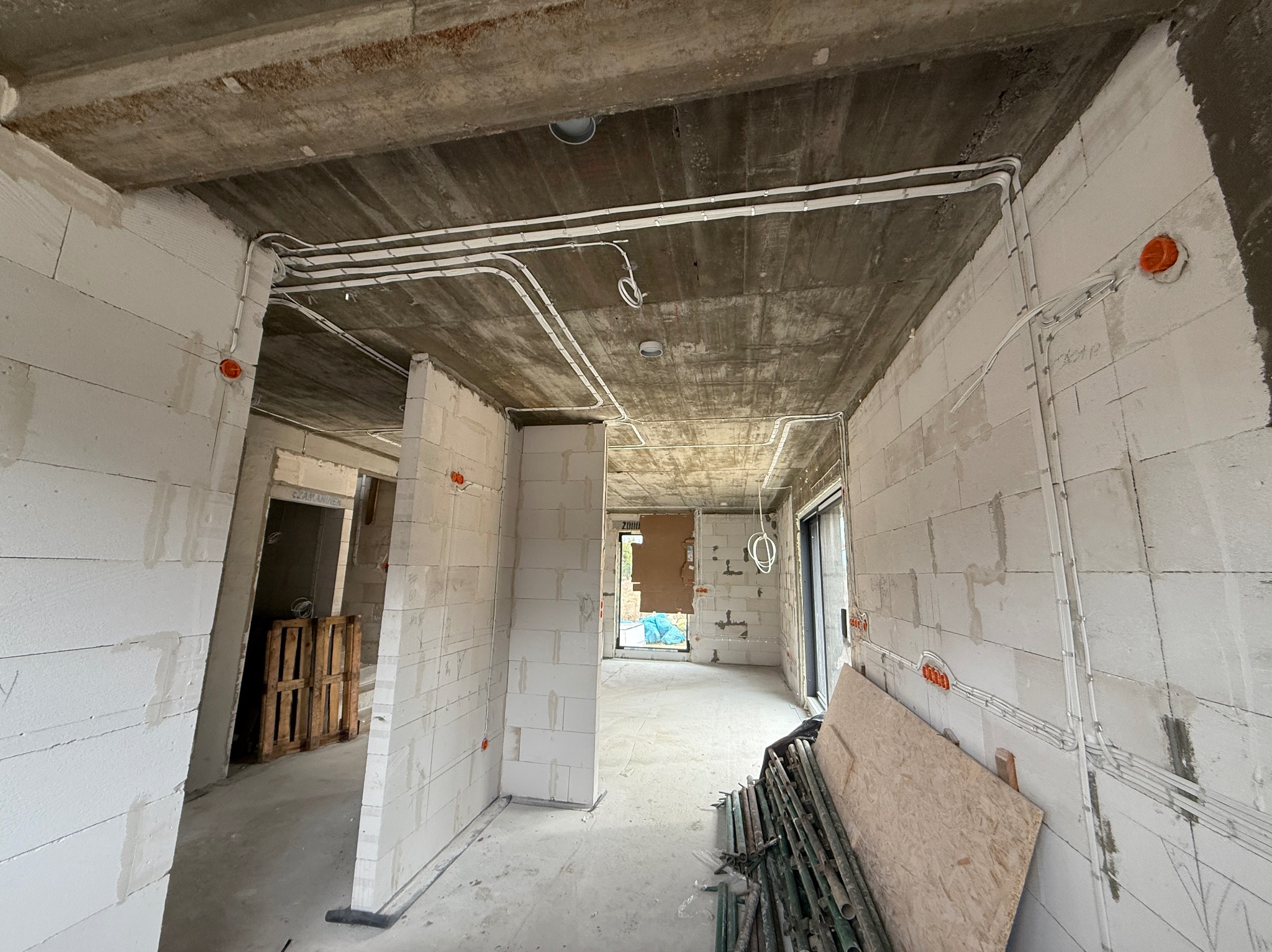 Surowy stan budynku z widoczną instalacją elektryczną: białe kable prowadzone po ścianach z bloków i pod sufitem. Widoczne puszki elektryczne i elementy konstrukcyjne.