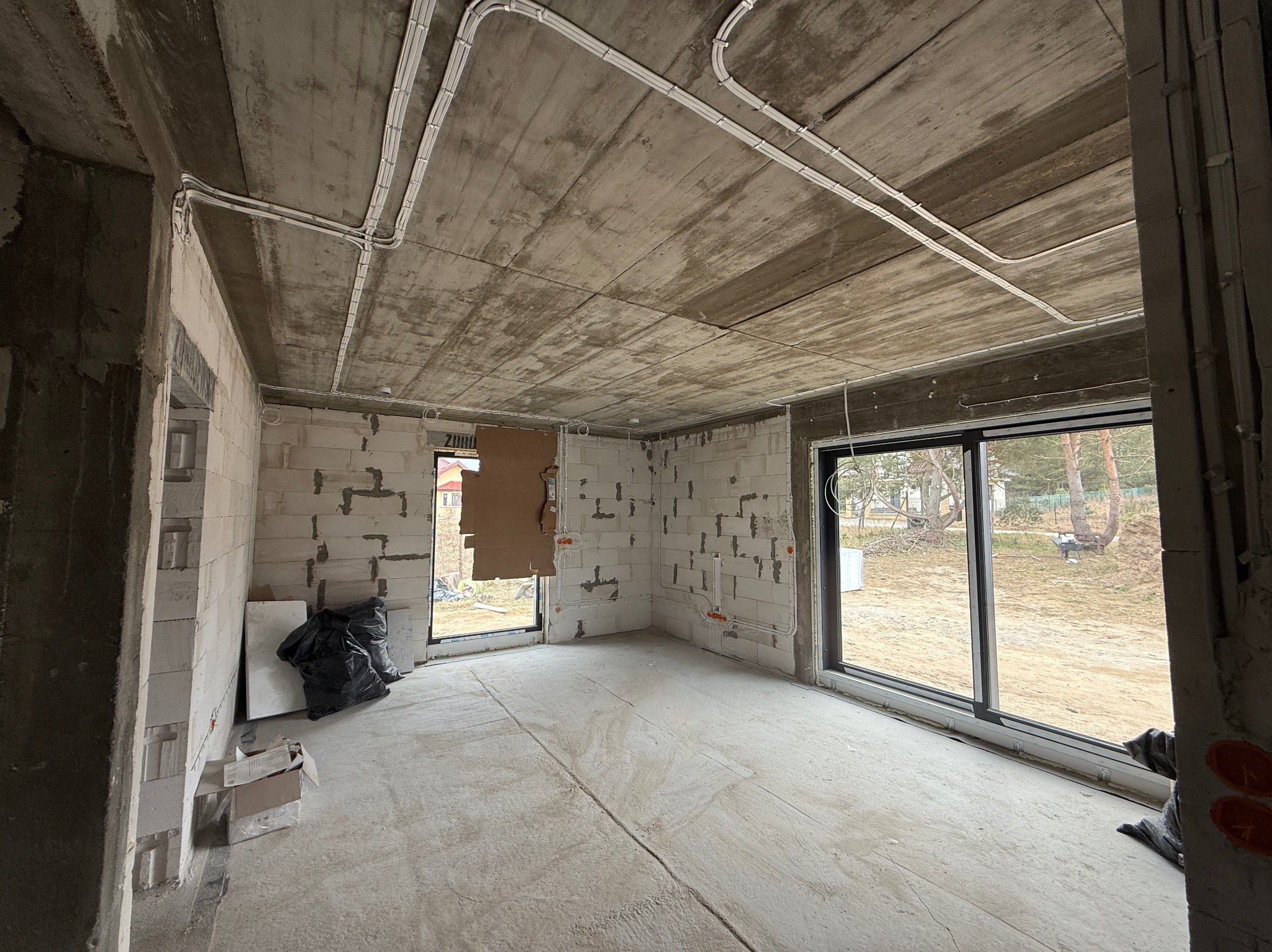 Wnętrze w stanie surowym z widoczną instalacją elektryczną w białych rurkach na suficie. Ściany z białych bloczków, duże okna. Na podłodze ślady po wylewce.
