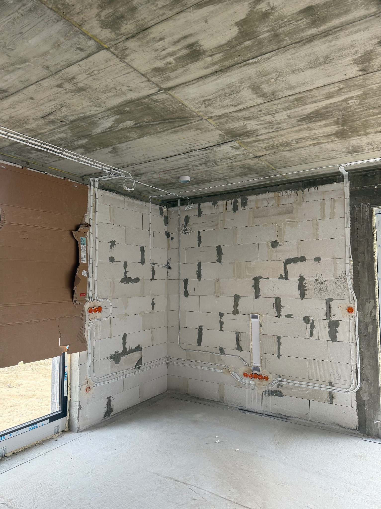 Surowy stan: instalacja elektryczna w nowym budynku z białego bloczka, widoczne kable i puszki, betonowy sufit, okno z kartonowym zabezpieczeniem.