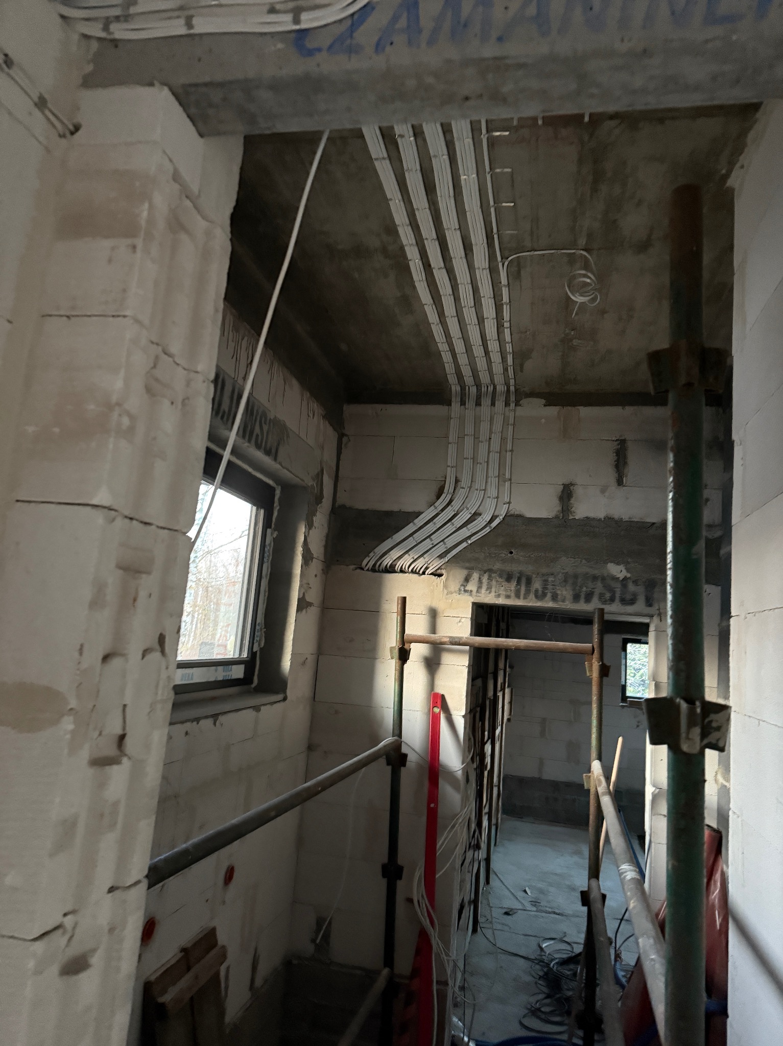 Instalacja elektryczna w surowym wnętrzu: kable ułożone w korytkach na suficie, ściany z betonu komórkowego, rusztowania. Widok na okno i dalszą część budynku.