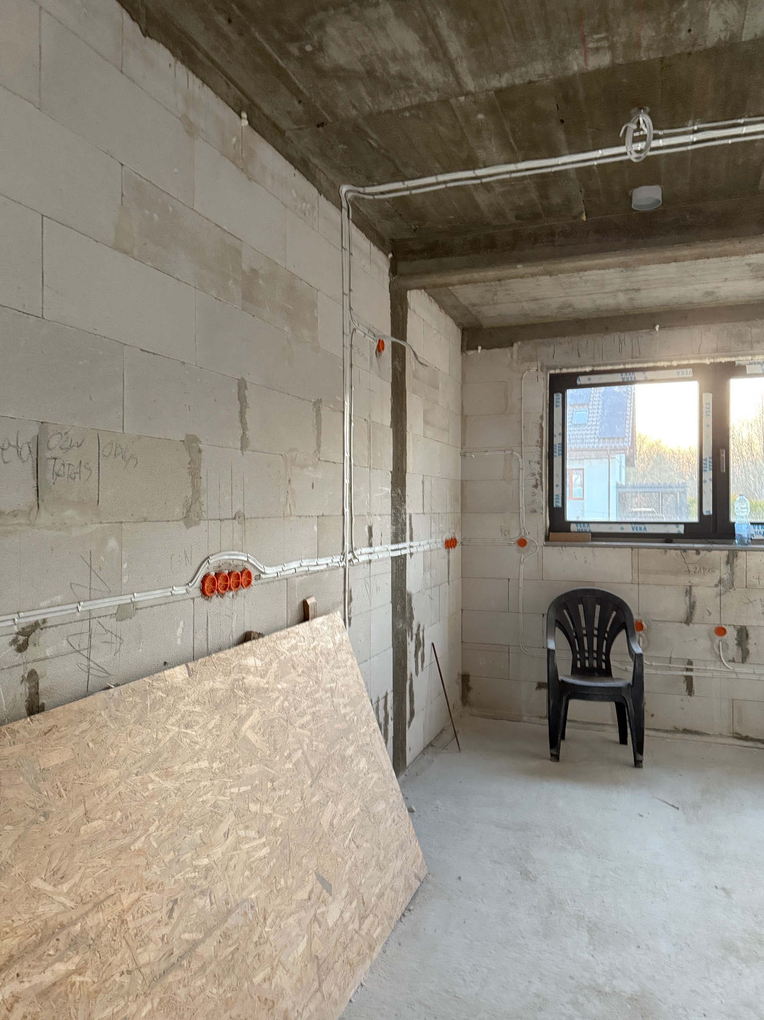Surowy stan: białe ściany z betonu komórkowego, widoczne przewody elektryczne w peszlach i pomarańczowe puszki. Na podłodze płyta OSB, czarne krzesło. Okno z widokiem na dom.