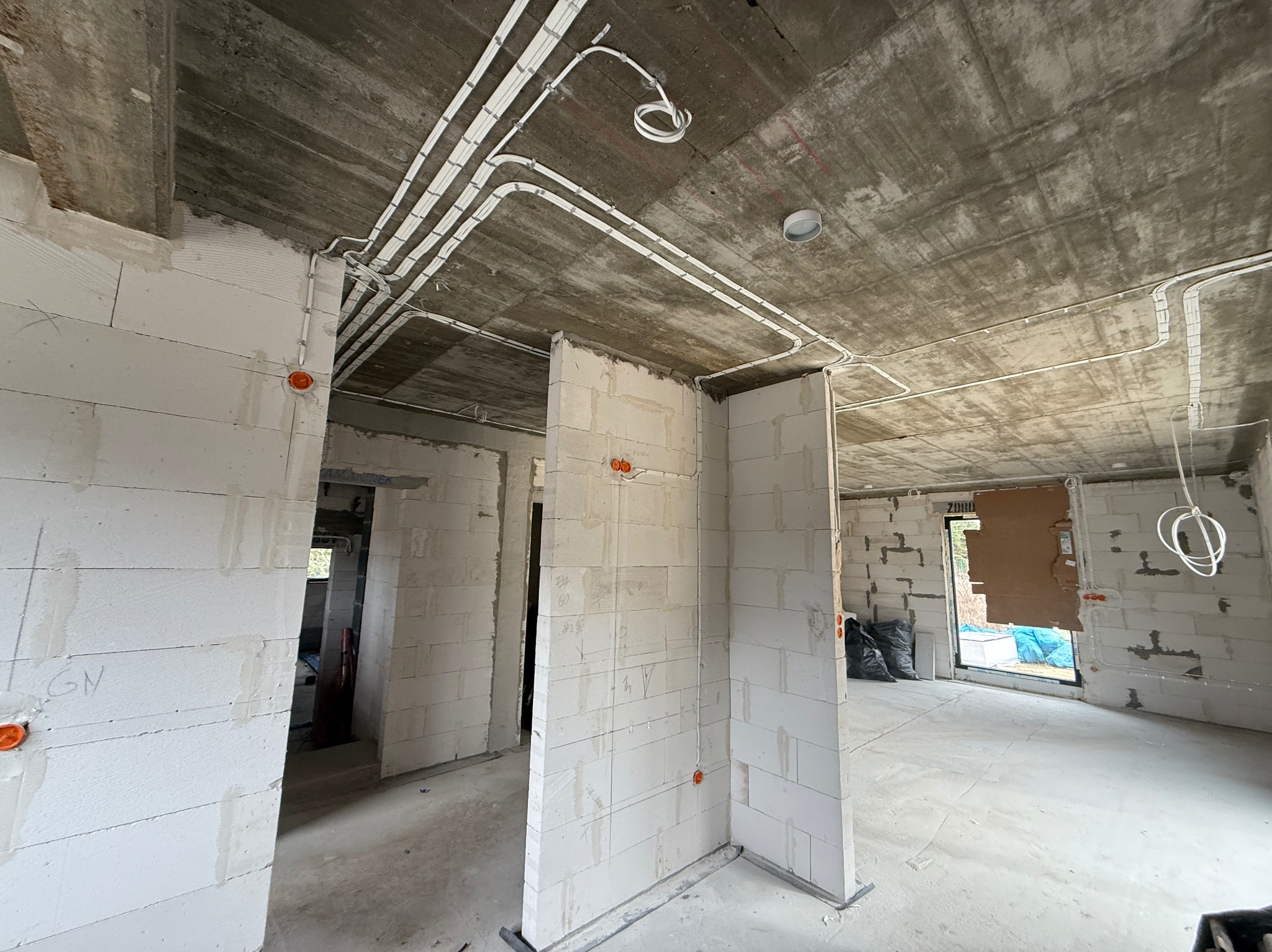 Surowy stan: białe ściany z betonu komórkowego, widoczne kable elektryczne prowadzone po suficie i ścianach, pomarańczowe puszki instalacyjne. Wnętrze w trakcie prac.