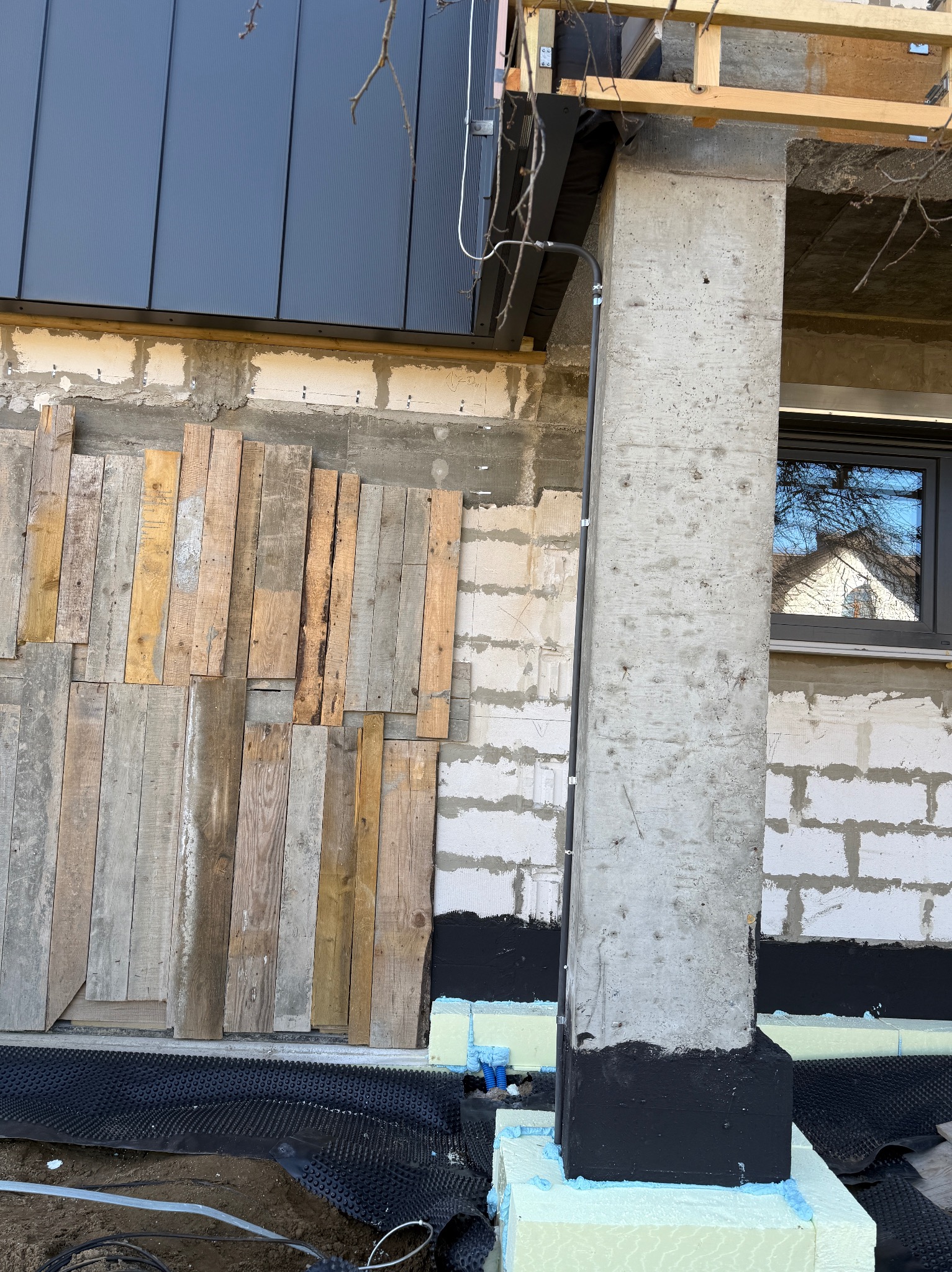 Surowy betonowy filar domu w trakcie budowy, z widoczną instalacją elektryczną w rurce, izolacją fundamentów i częściowo ocieploną ścianą z drewnianym szalunkiem.