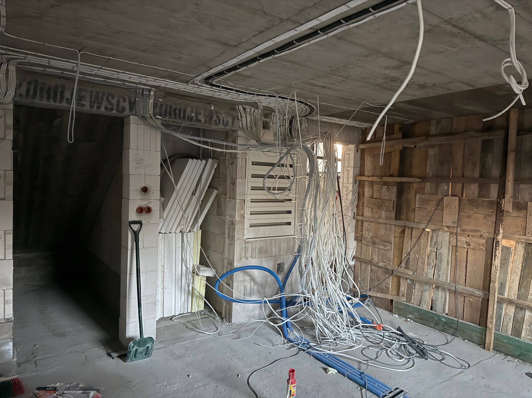 Surowy stan instalacji elektrycznej i hydraulicznej w trakcie budowy: kable, rury, szpadel, betonowy strop z widocznymi przewodami. Wnętrze w stanie deweloperskim.