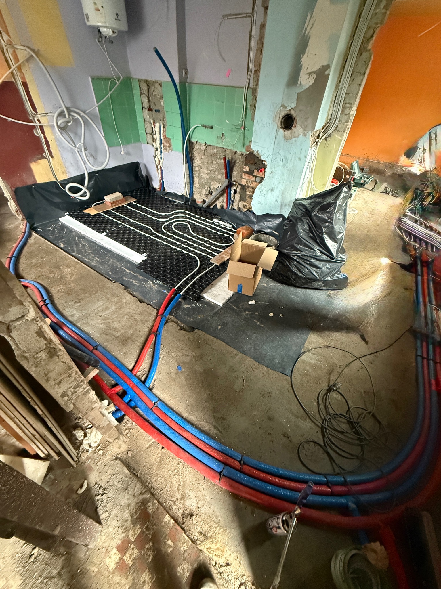 Montaż ogrzewania podłogowego: białe rurki ułożone na czarnej macie, czerwono-niebieskie rury doprowadzające, widoczne elementy instalacji w surowym wnętrzu.