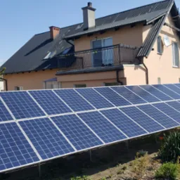 Instalacja paneli słonecznych na gruncie przed domem jednorodzinnym z czarnym dachem i balkonem.