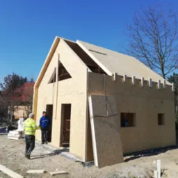 Wznoszony dom w konstrukcji szkieletowej z płyt OSB, widoczni dwaj pracownicy w ubraniach roboczych, niebieskie niebo w tle.