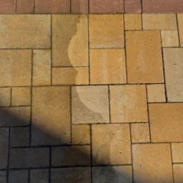 Fragment powierzchni z kostki brukowej w odcieniach beżu i brązu, z widocznym cieniem i śladami wilgoci po niedawnym czyszczeniu.