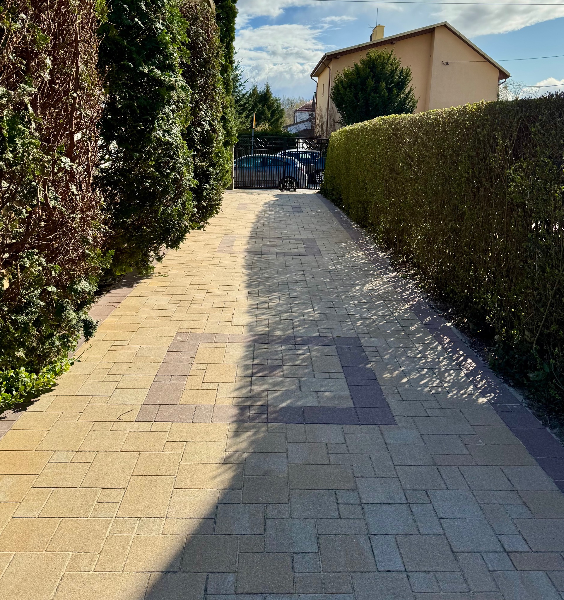 Ułożona z kostki brukowej ścieżka prowadząca do bramy wjazdowej, otoczona równo przyciętymi żywopłotami, z widocznym samochodem za bramą.