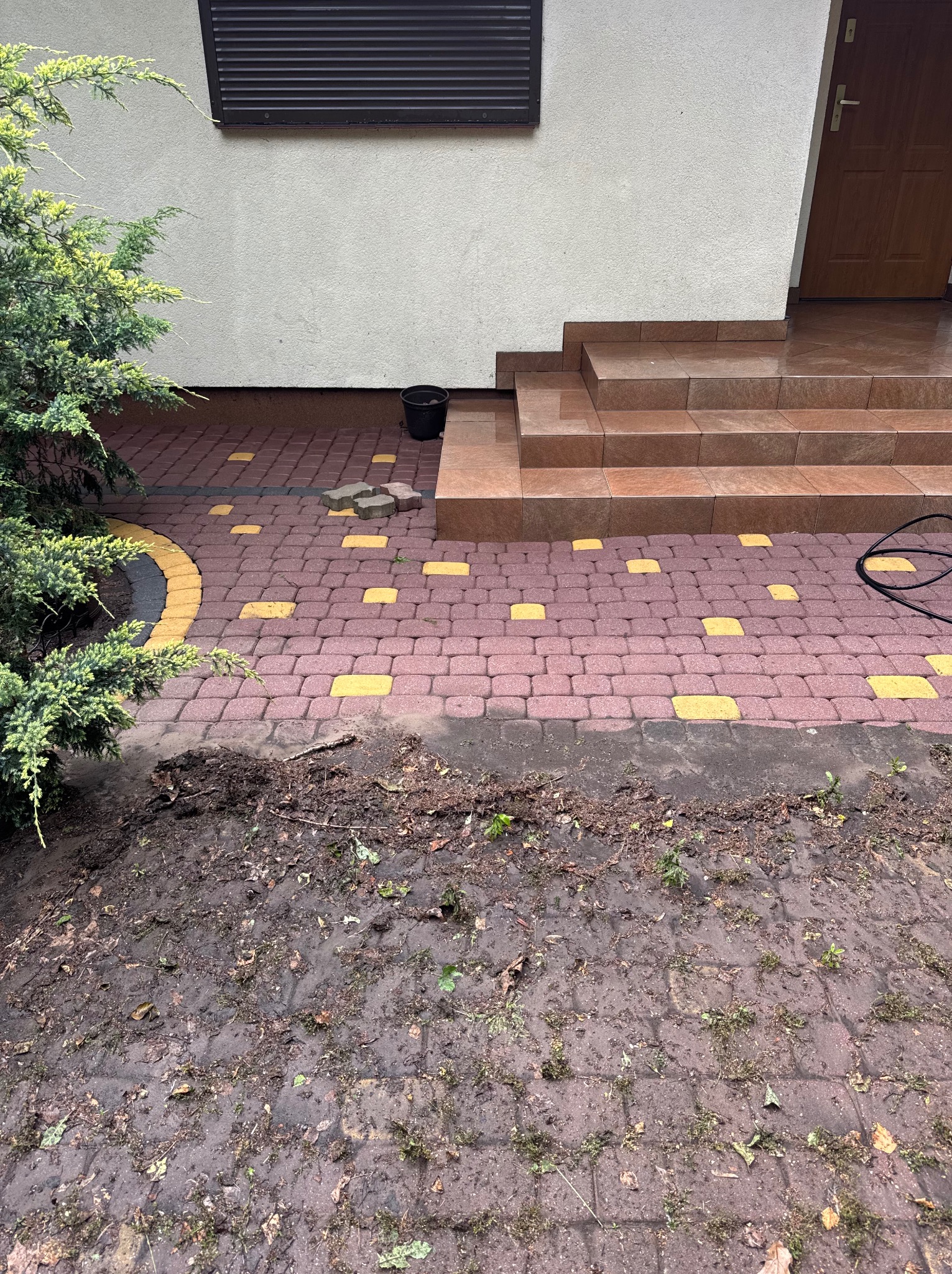 Ułożona kostka brukowa w odcieniach czerwieni i żółci przed domem, część powierzchni zabrudzona ziemią, obok rosnący krzew. Widoczne schody wejściowe.