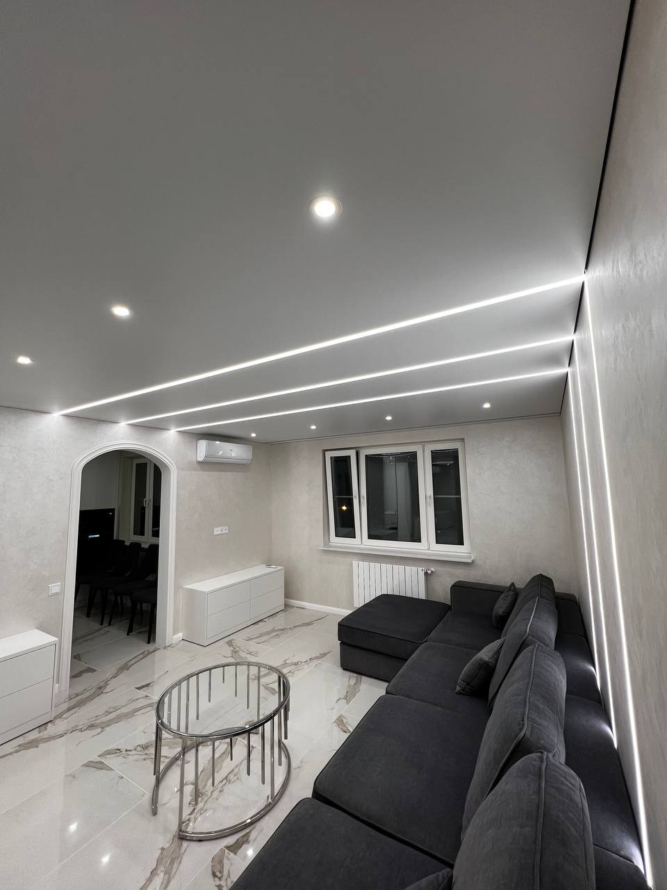 Nowoczesny salon z szarym narożnikiem, marmurową podłogą i oświetleniem LED w suficie podwieszanym.