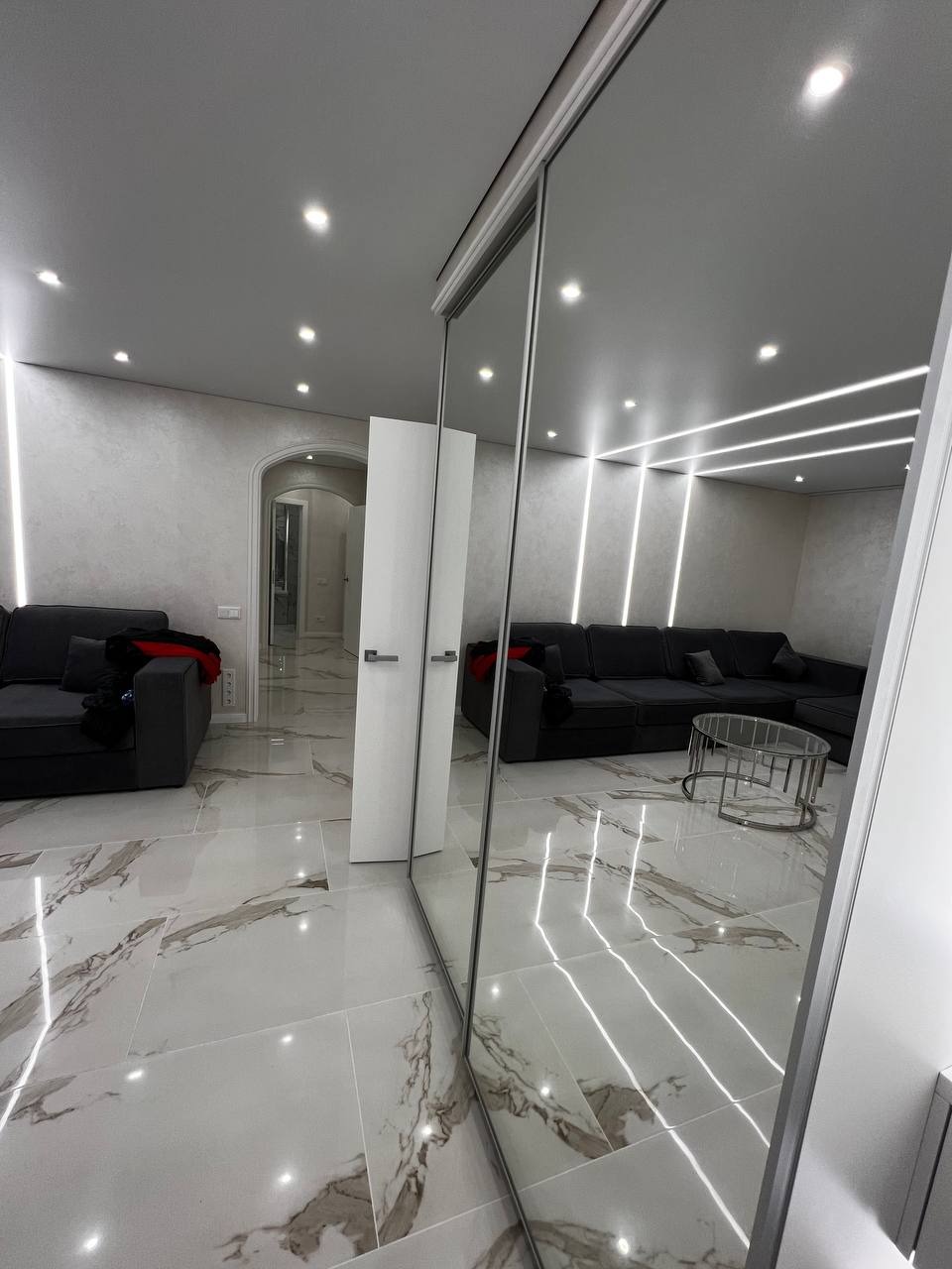 Nowoczesne wnętrze salonu z marmurową podłogą, szarą sofą narożną i lustrzaną szafą przesuwną, oświetlone ledowymi liniami na suficie.