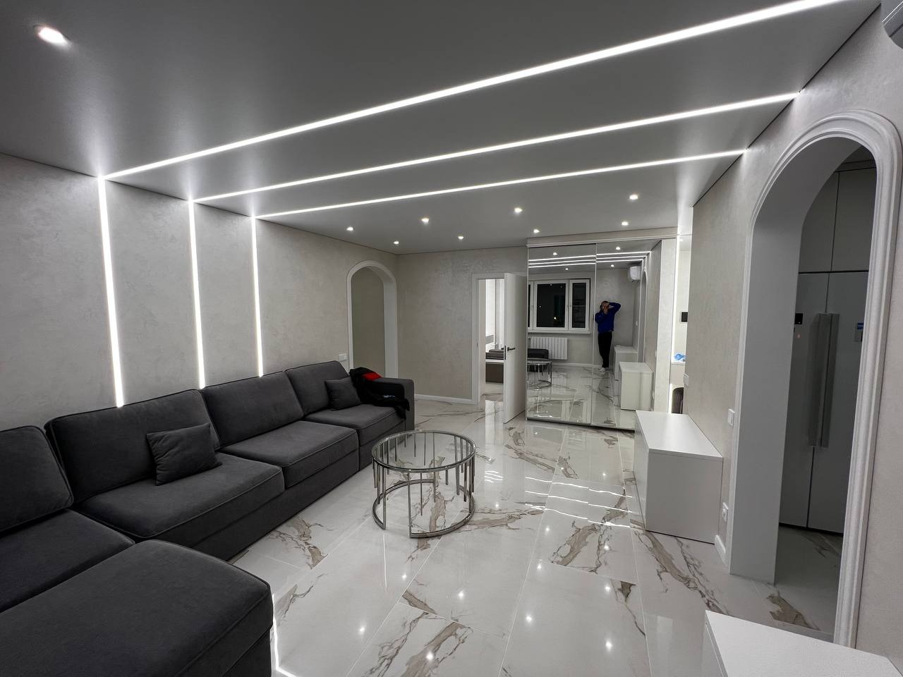 Nowoczesny salon z szarą sofą narożną, okrągłym szklanym stolikiem kawowym, marmurową podłogą i oświetleniem LED w suficie oraz ścianach.