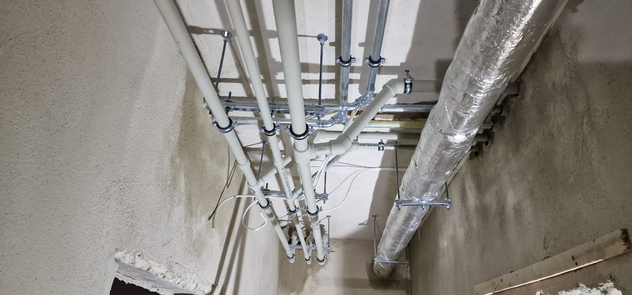 Widok z dołu na zamontowane rury instalacji wodnej i wentylacyjnej pod sufitem w surowym stanie, widoczne mocowania i izolacja termiczna kanału wentylacyjnego.