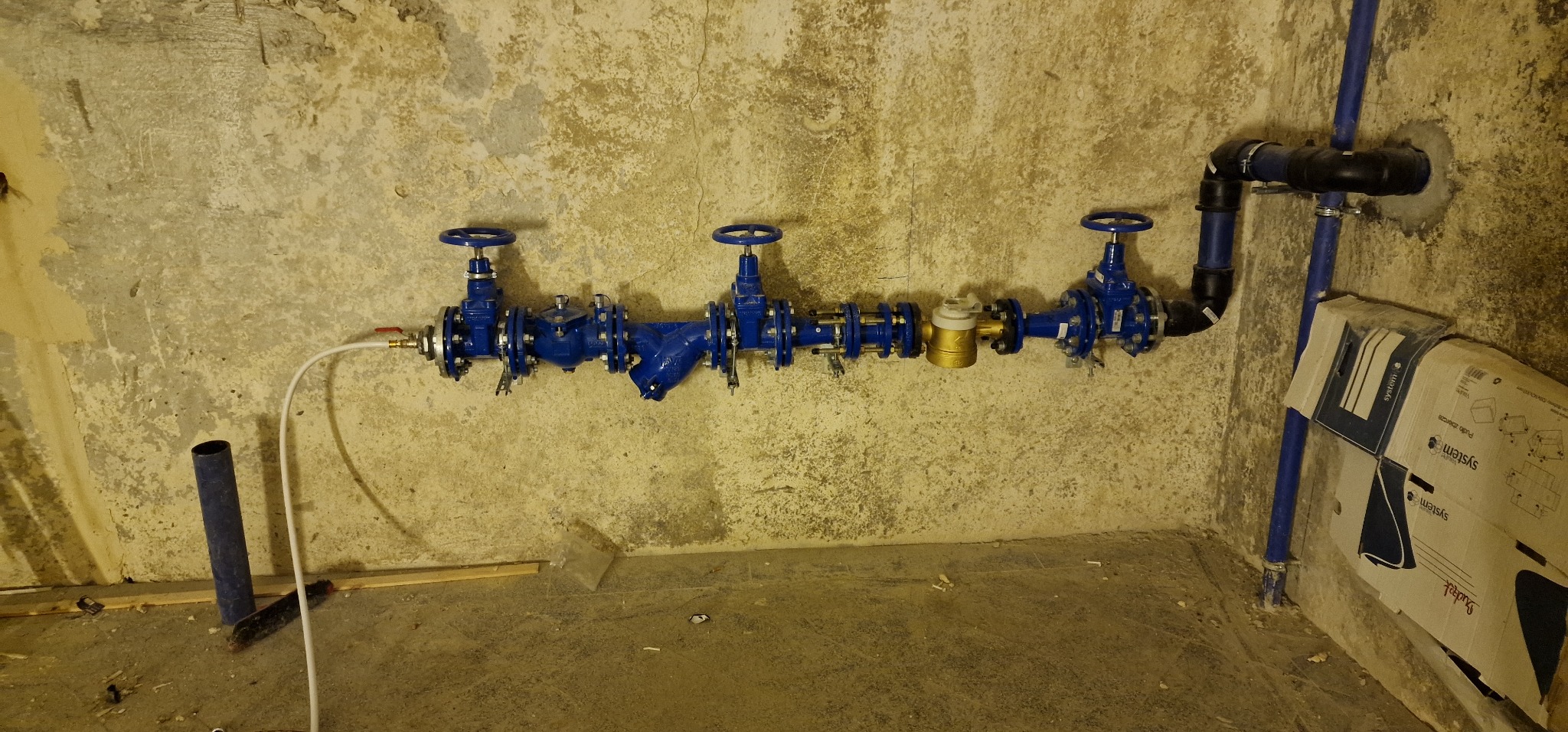 Niebieska instalacja wodna z zaworami i licznikiem, zamontowana na surowej ścianie w pomieszczeniu w trakcie prac instalacyjnych, widoczny karton z opisem systemu.