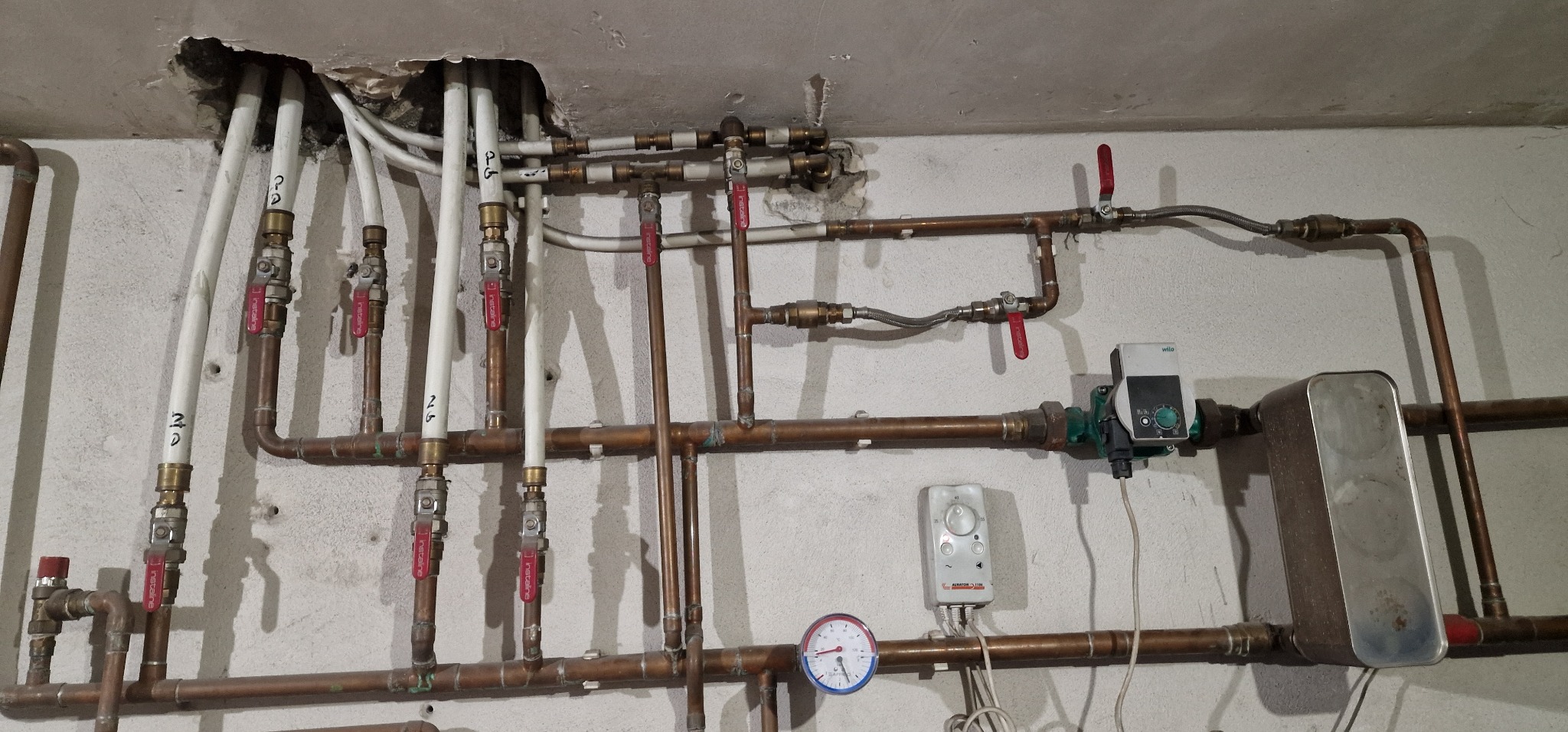 Miedziane i białe rury instalacji grzewczej z zaworami, manometrem, pompą obiegową i sterownikiem, zamontowane na szarej ścianie z widocznymi śladami po kuciu w stropie.