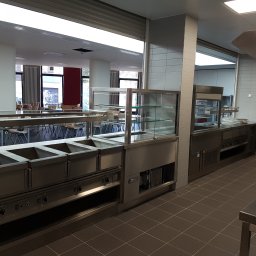 SIBI1.PL SP. Z O.O. - Wnętrze stołówki z profesjonalnym wyposażeniem kuchennym ze stali nierdzewnej: bemary, witryna chłodnicza, stanowisko do wydawania posiłków, widoczne stoliki i krzesła dla gości.