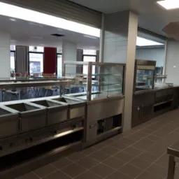 Wnętrze stołówki z profesjonalnym wyposażeniem kuchennym ze stali nierdzewnej: bemary, witryna chłodnicza, stanowisko do wydawania posiłków, widoczne stoliki i krzesła dla gości.