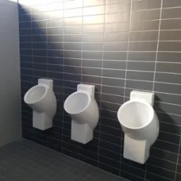 Trzy białe pisuary zamontowane na ścianie wyłożonej szarymi płytkami w publicznej toalecie, z widocznym grzejnikiem na ścianie bocznej.