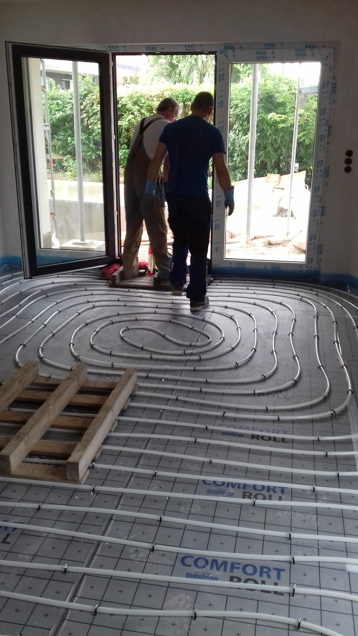Montaż podłogowego ogrzewania wodnego: widok na rozłożone rury grzewcze na izolacji, z pracownikami w tle przy oknie balkonowym. Paleta drewniana leży na podłodze.