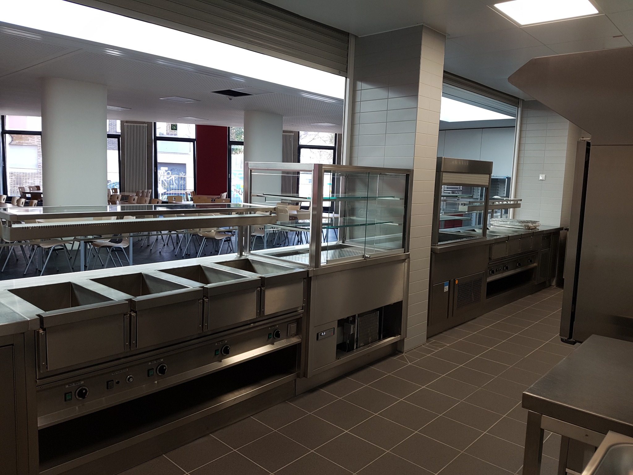 Wnętrze stołówki z profesjonalnym wyposażeniem kuchennym ze stali nierdzewnej: bemary, witryna chłodnicza, stanowisko do wydawania posiłków, widoczne stoliki i krzesła dla gości.