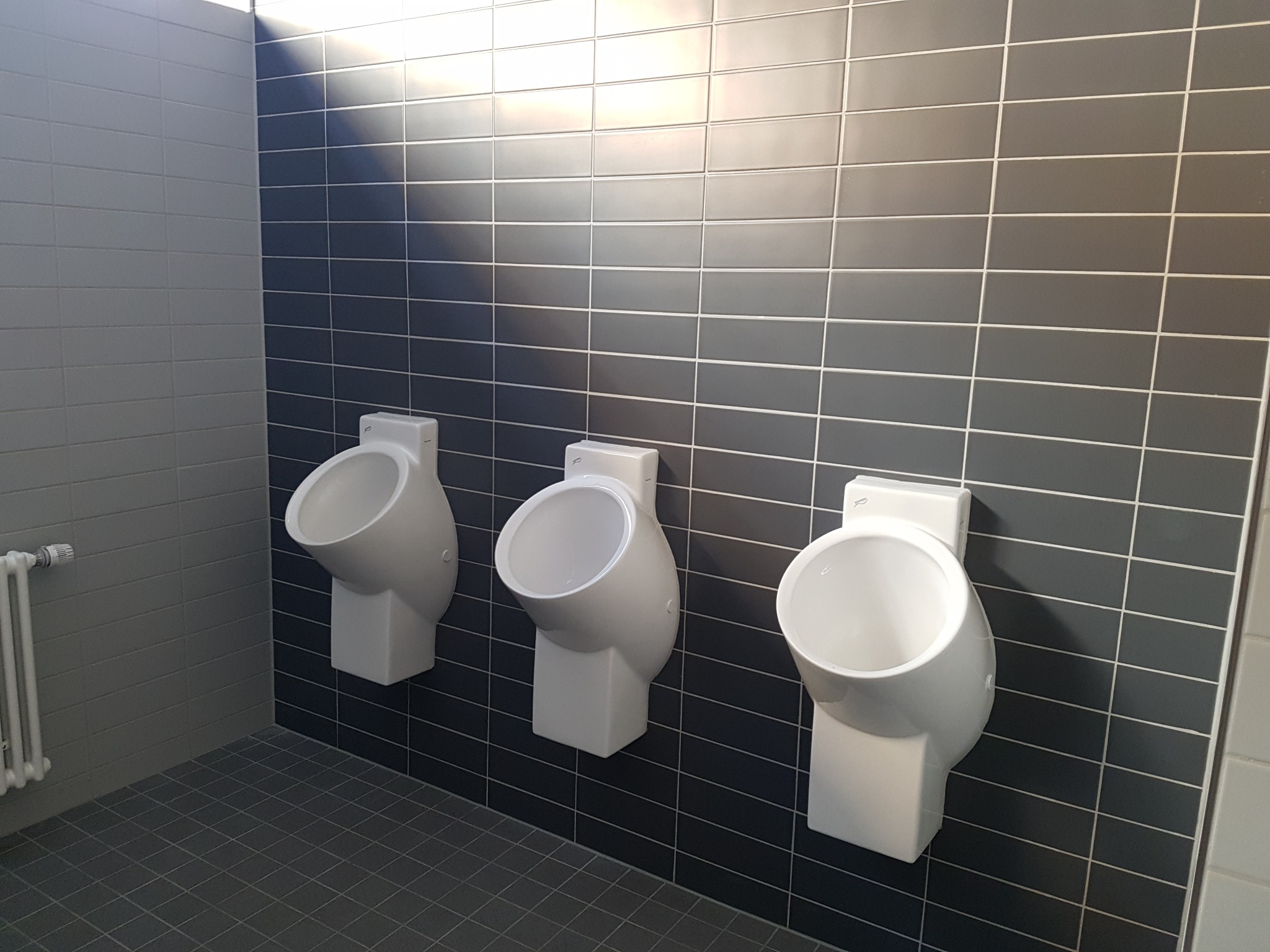 Trzy białe pisuary zamontowane na ścianie wyłożonej szarymi płytkami w publicznej toalecie, z widocznym grzejnikiem na ścianie bocznej.