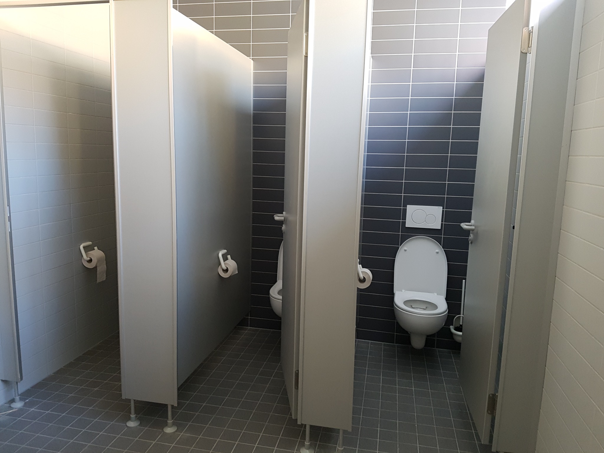 Publiczna toaleta z kabinami oddzielonymi białymi ściankami, widoczne muszle klozetowe i uchwyty na papier toaletowy, szare płytki na ścianach i podłodze.