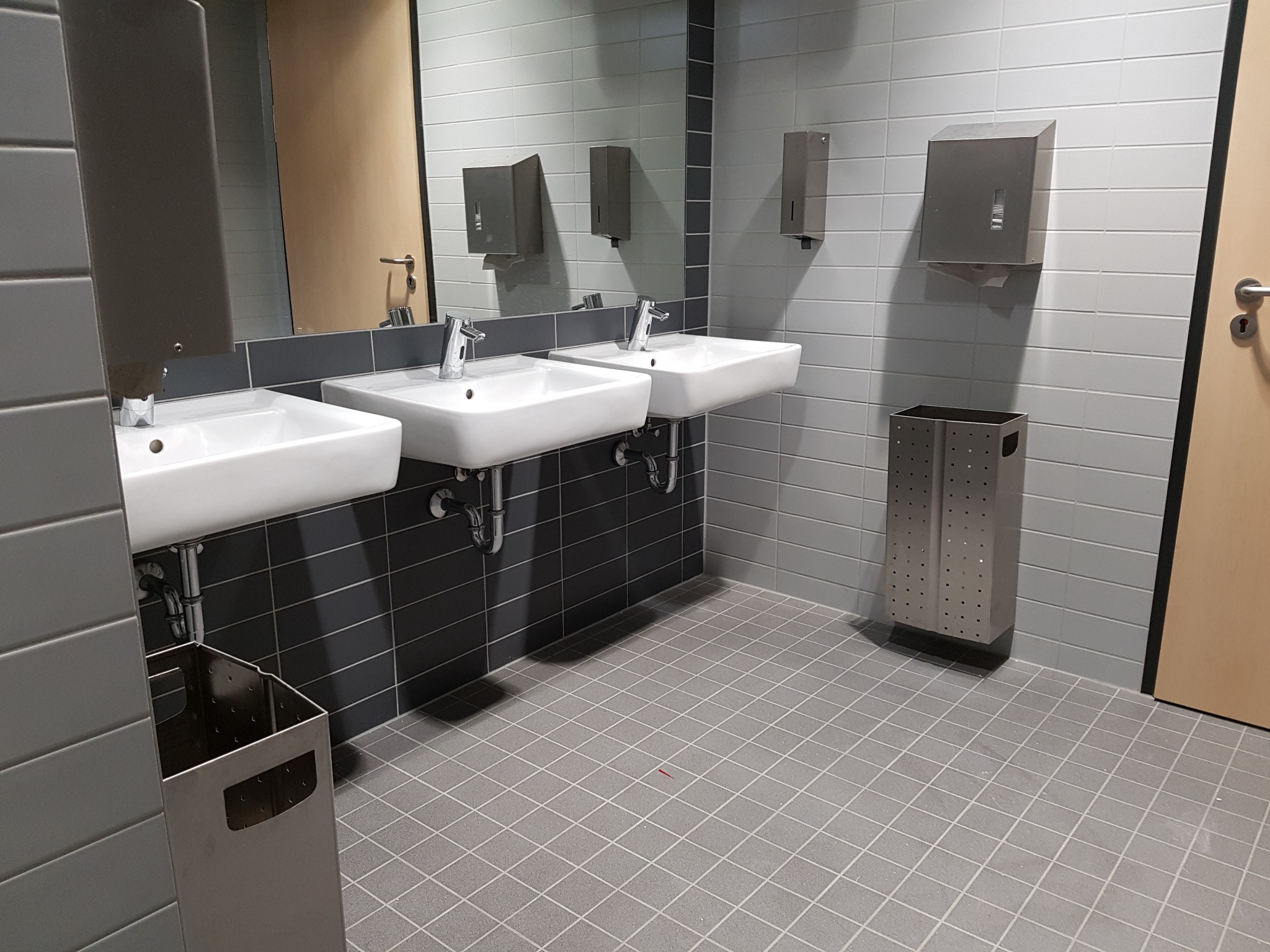 Publiczna toaleta z trzema umywalkami, automatycznymi kranami, dozownikami mydła i ręczników, oraz koszami na śmieci, wykończona szarymi płytkami.