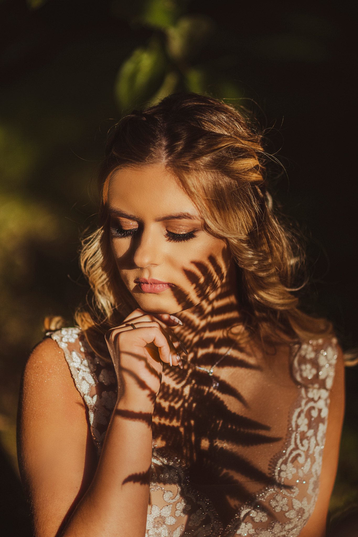 Panna młoda w sukni ślubnej, z delikatnym makijażem, zamyślona, oświetlona promieniami słońca, z cieniem liścia paproci na twarzy.