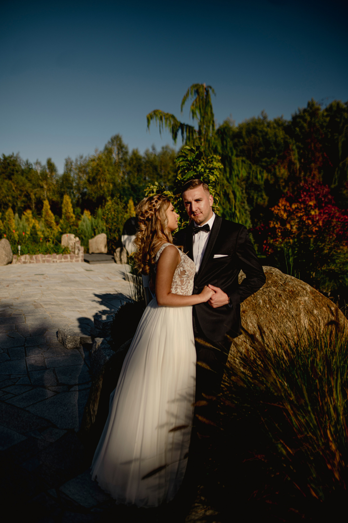 Para młoda w ogrodzie: Panna młoda w sukni z koronkową górą i wiankiem we włosach trzyma za rękę pana młodego w smokingu, opierając się o duży kamień na tle zieleni i kamiennej ścieżki.