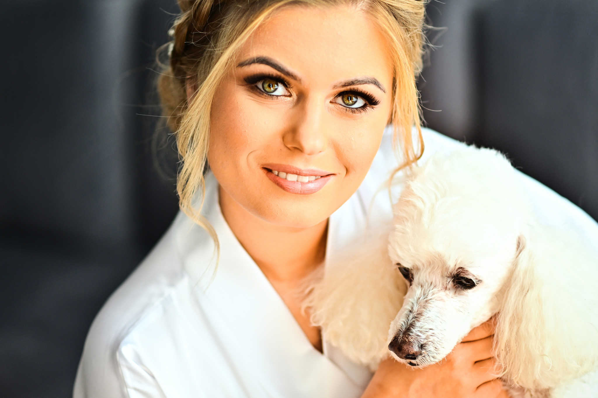 Portret uśmiechniętej blondynki w białym szlafroku, trzymającej białego pudla na tle szarej ściany.