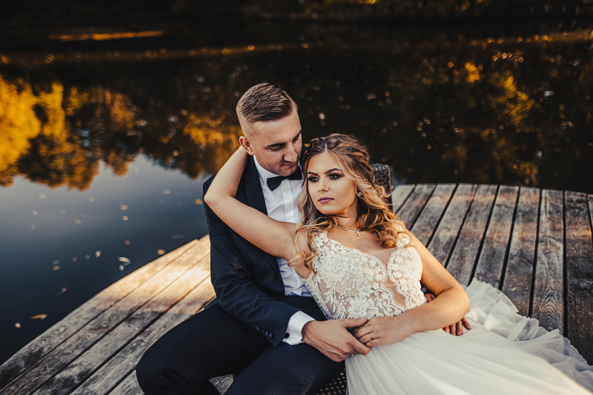 Para młoda siedzi na drewnianym pomoście nad jeziorem, panna młoda w koronkowej sukni z odkrytymi ramionami i delikatną biżuterią, pan młody w czarnym garniturze z muchą, jesienna sceneria w tle.