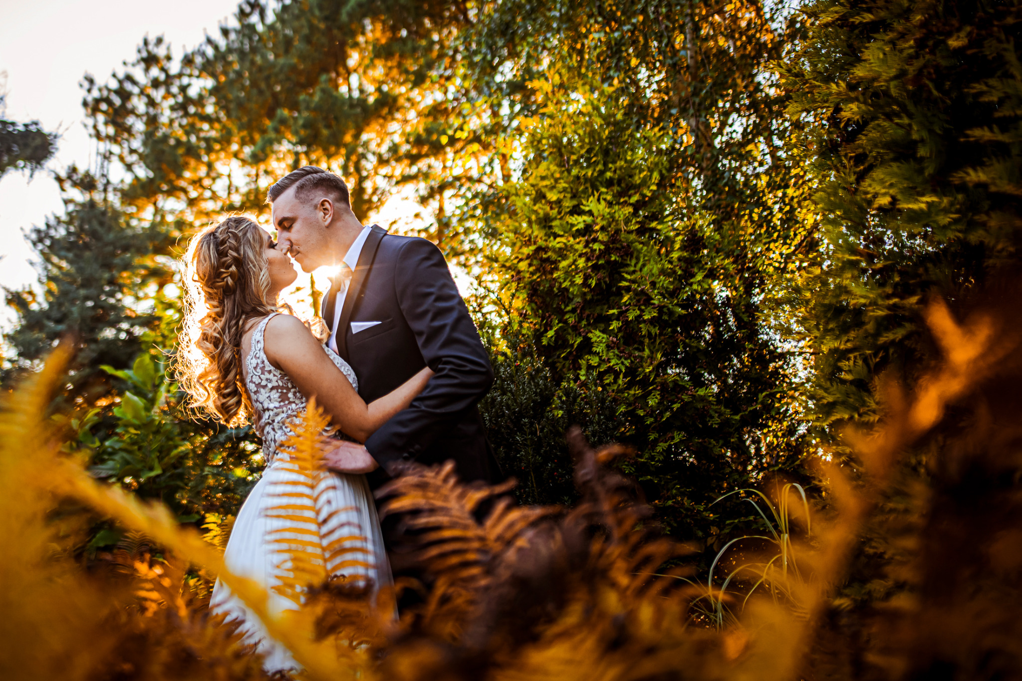 Para młoda w objęciach, pocałunek na tle zieleni i promieni słonecznych, panna młoda w sukni z koronkową górą i upiętymi włosami.