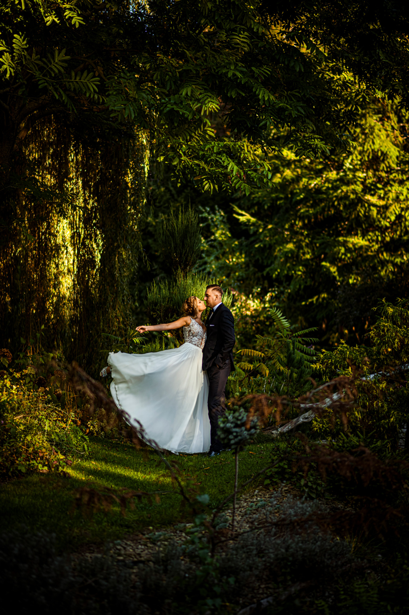 Para młoda w leśnej scenerii: Panna młoda w koronkowej sukni z rozkloszowanym dołem, unoszona przez pana młodego, pocałunek na tle drzew i krzewów.