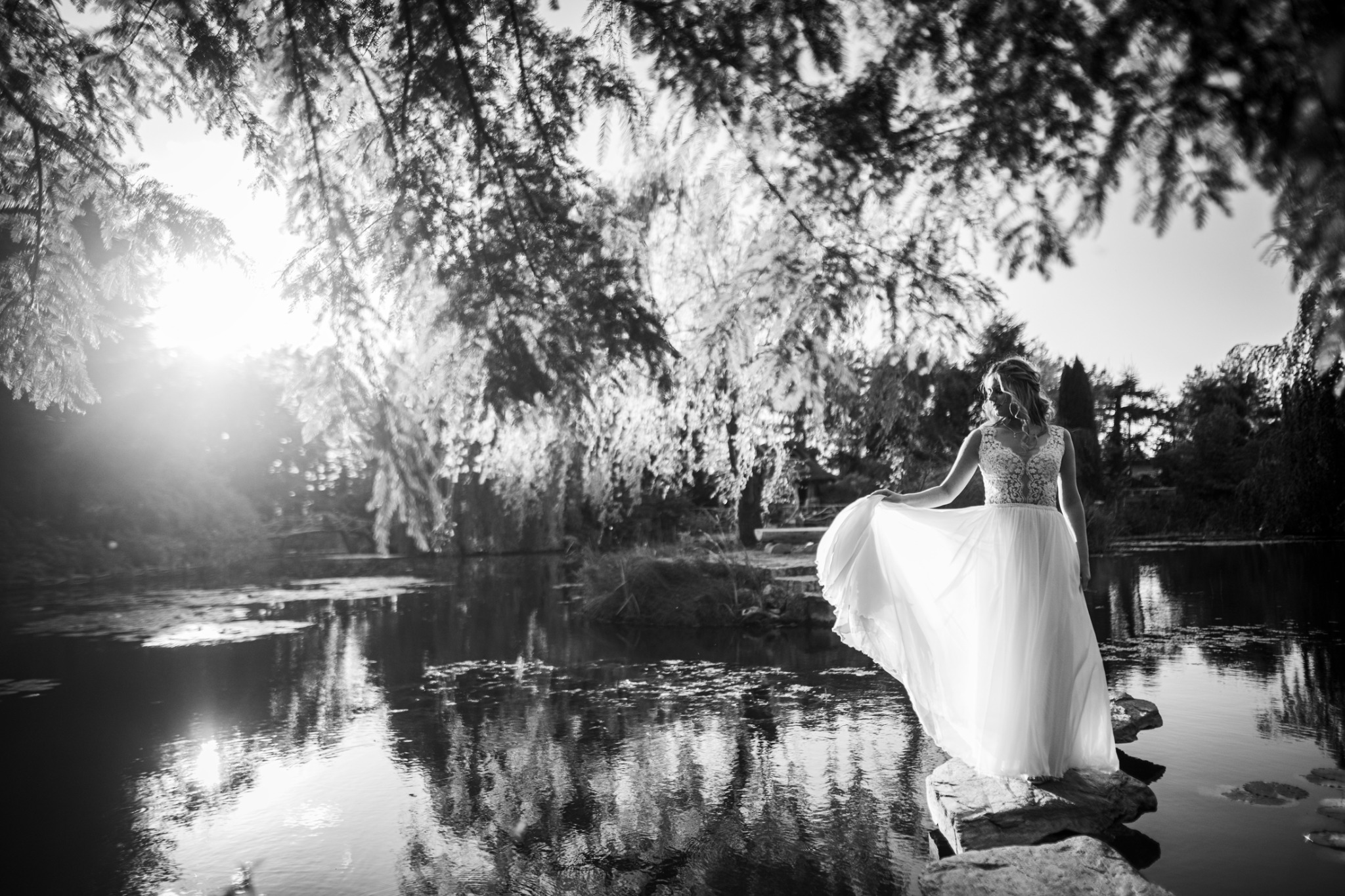 Panna młoda w koronkowej sukni, stojąca na kamieniu nad wodą, w czarno-białej scenerii z refleksami światła w wodzie i zwisającymi gałęziami drzew
