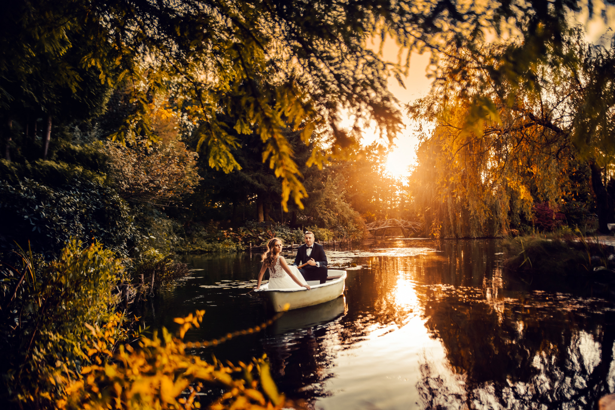 Para młoda w łódce na jeziorze o zachodzie słońca, oboje elegancko ubrani, ona w białej sukni, on w smokingu, otoczeni drzewami z jesiennymi liśćmi.