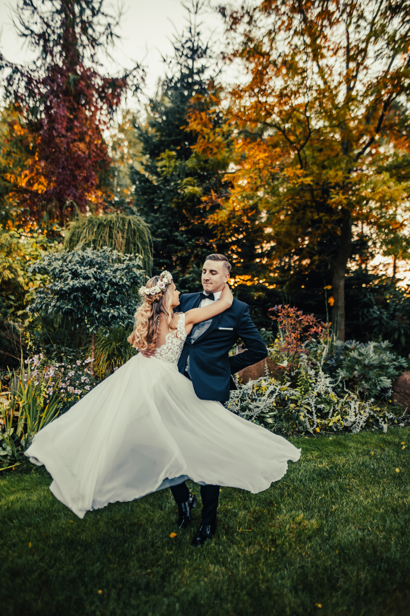 Para młoda tańczy w ogrodzie jesienią, panna młoda w koronie z kwiatów i zwiewnej sukni, pan młody w eleganckim smokingu.