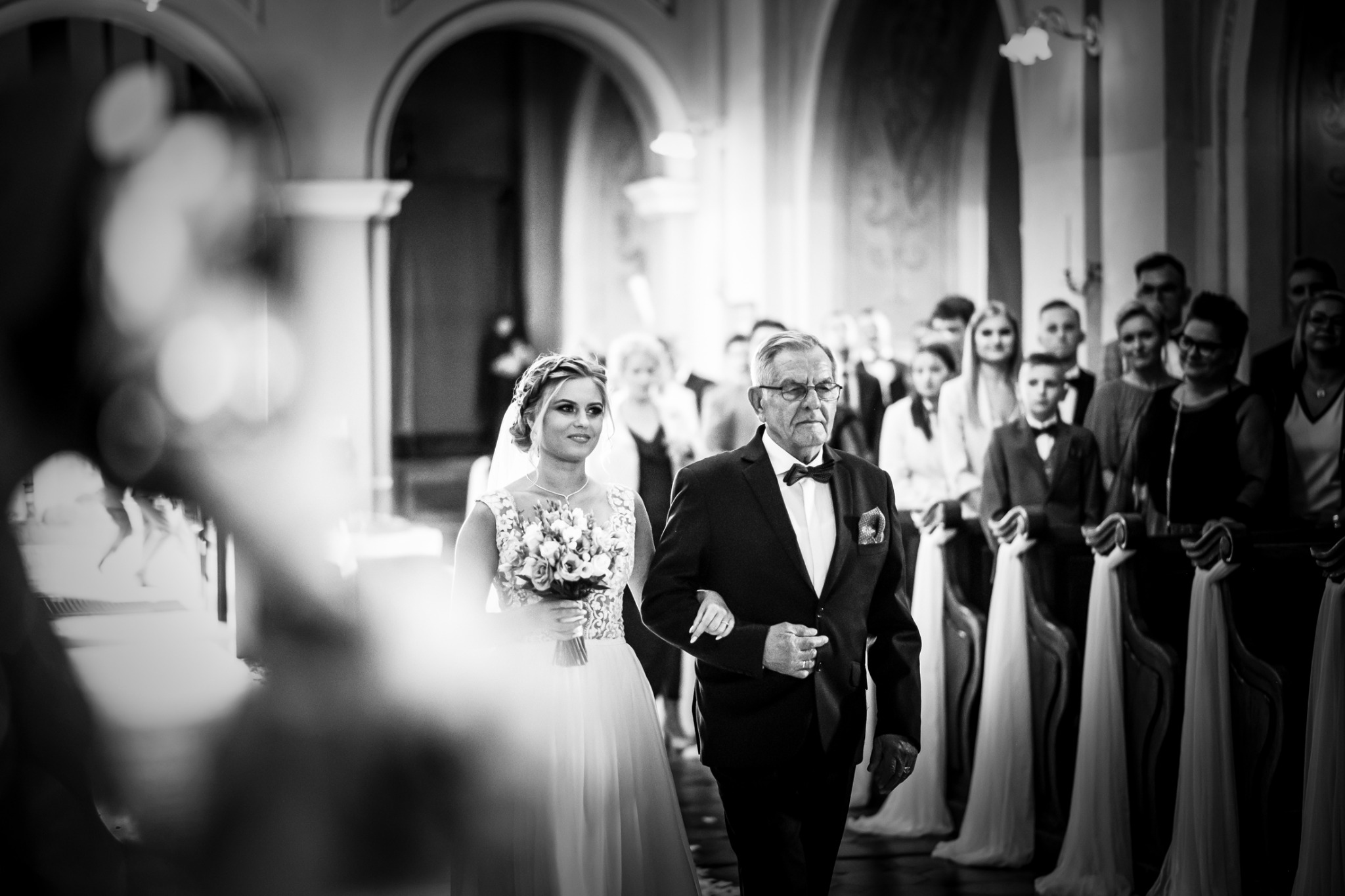 Czarno-białe zdjęcie panny młodej prowadzonej do ołtarza przez ojca, w tle widoczni goście weselni w kościele.