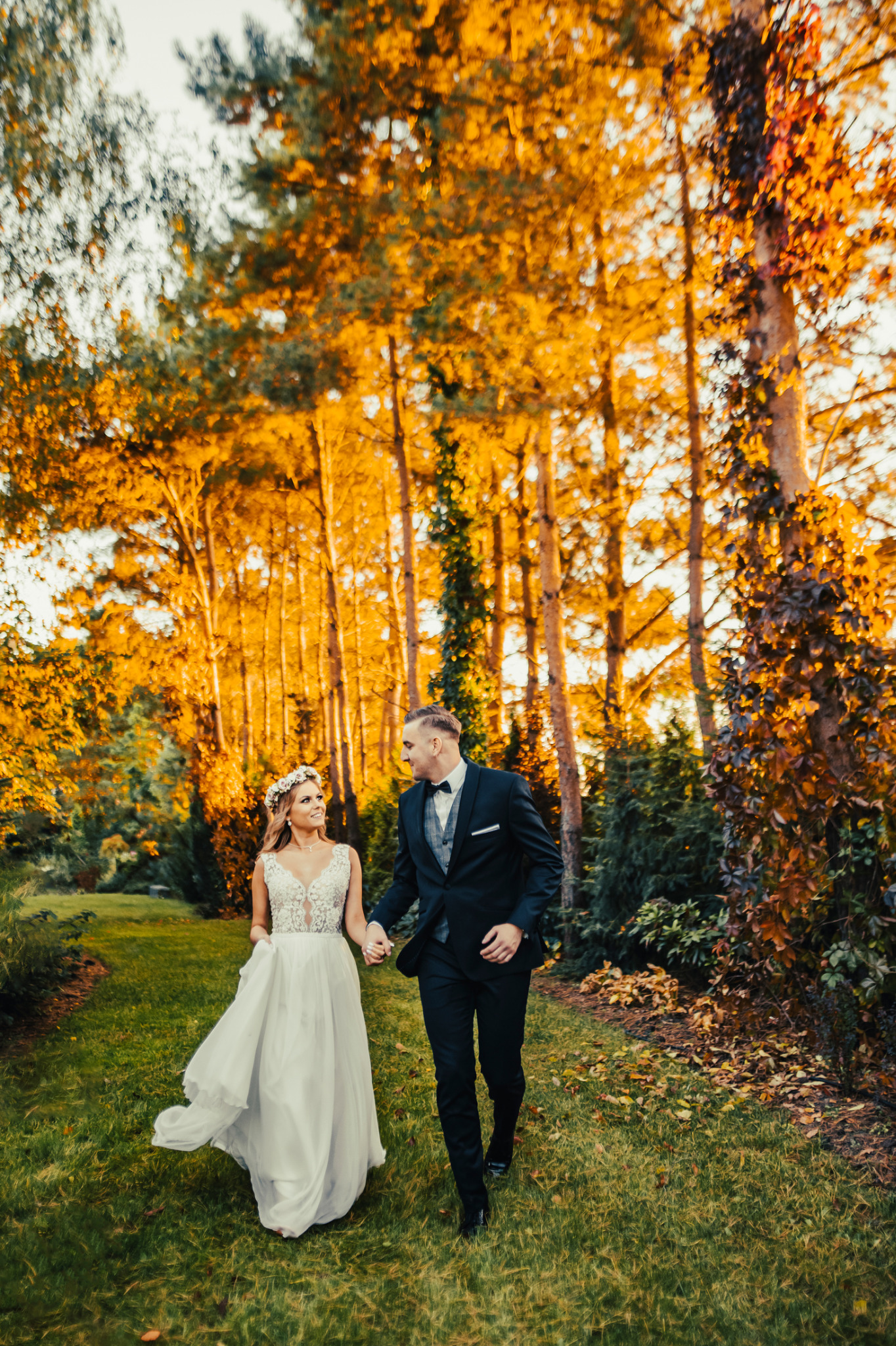 Uśmiechnięta panna młoda w koronkowej sukni i wianku, trzymająca za rękę eleganckiego pana młodego w czarnym smokingu, spacerują po zielonym trawniku w otoczeniu jesiennych drzew z pomarańczowymi...