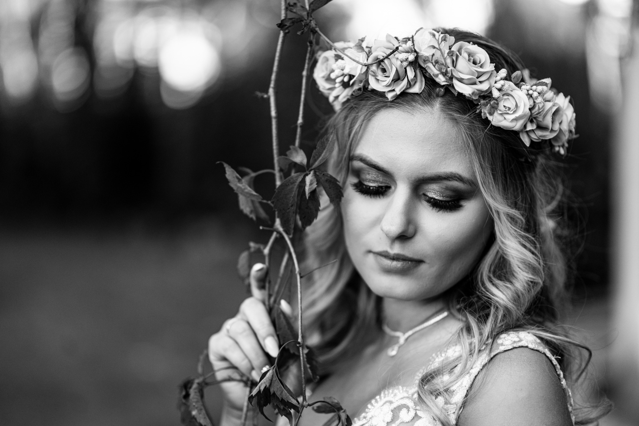 Czarno-białe zbliżenie panny młodej z wiankiem z róż na głowie, trzymającej gałązkę z liśćmi, skierowanej wzrokiem w dół, z delikatnym makijażem i biżuterią.