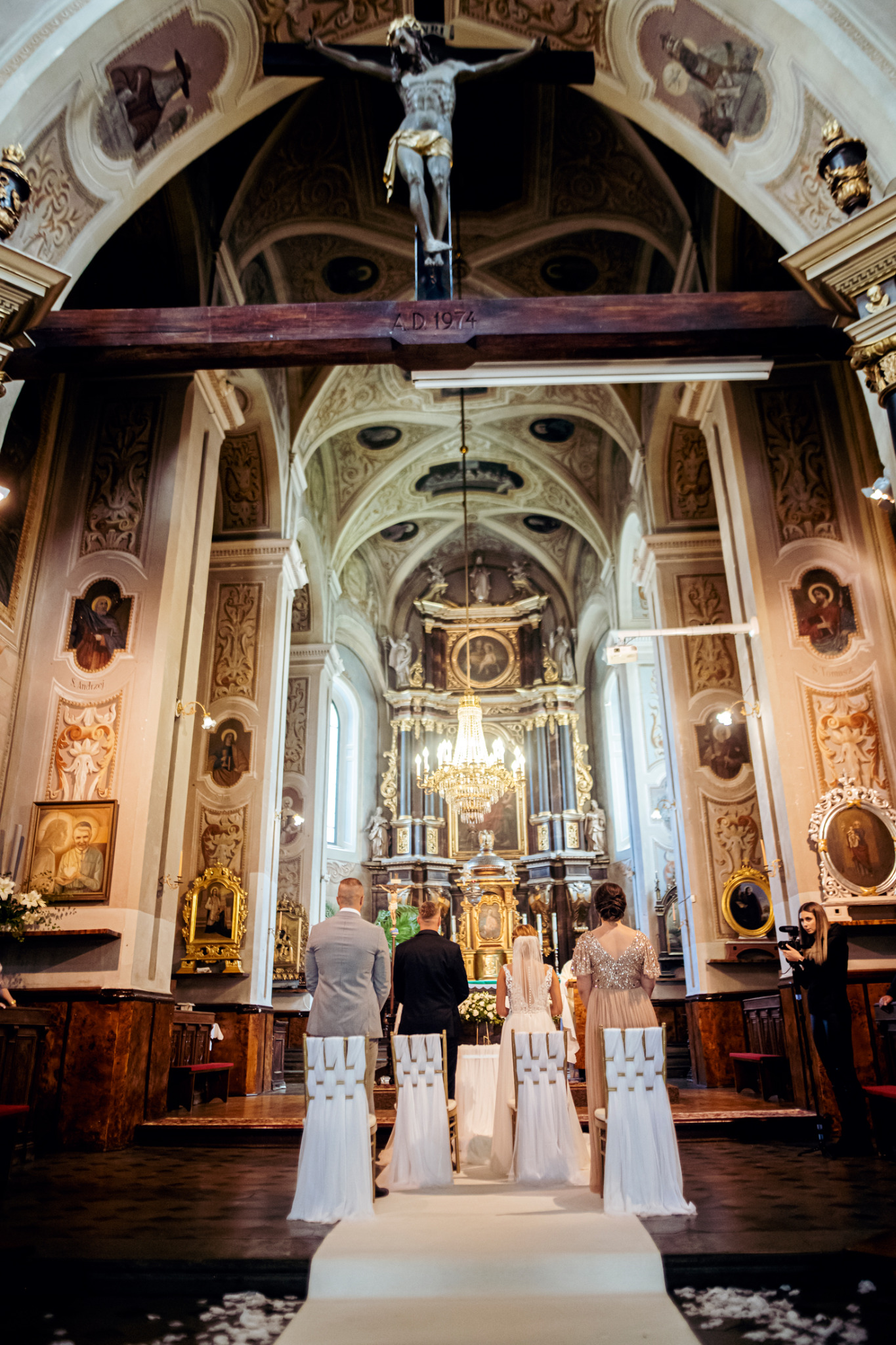 Para młoda podczas ceremonii ślubnej w bogato zdobionym kościele, widok zza udekorowanych krzeseł, z fotografem rejestrującym wydarzenie z boku.