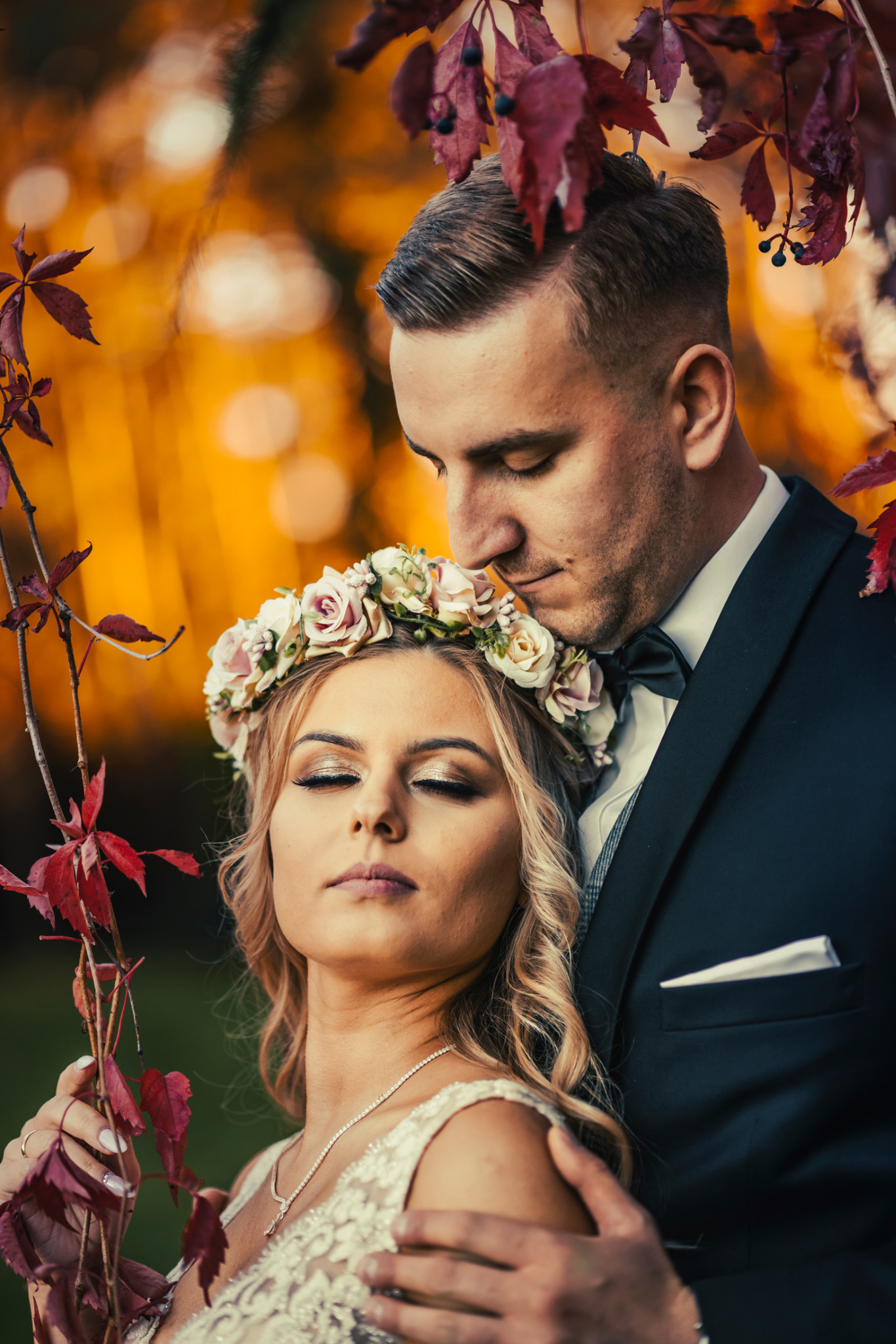 Elegancka para młoda w jesiennej scenerii: panna młoda z wiankiem z róż na głowie, pan młody w czarnym smokingu, delikatne oświetlenie i czerwone liście tworzą romantyczny nastrój.