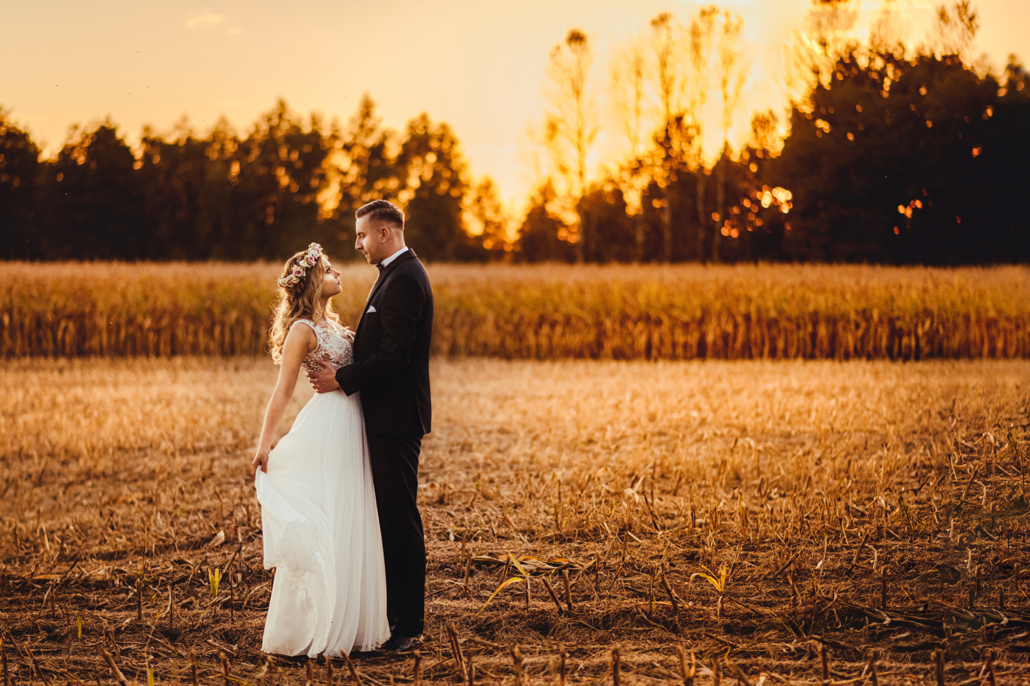 Para młoda w objęciach na polu ścierniska o zachodzie słońca, panna młoda w wianku i koronkowej sukni.