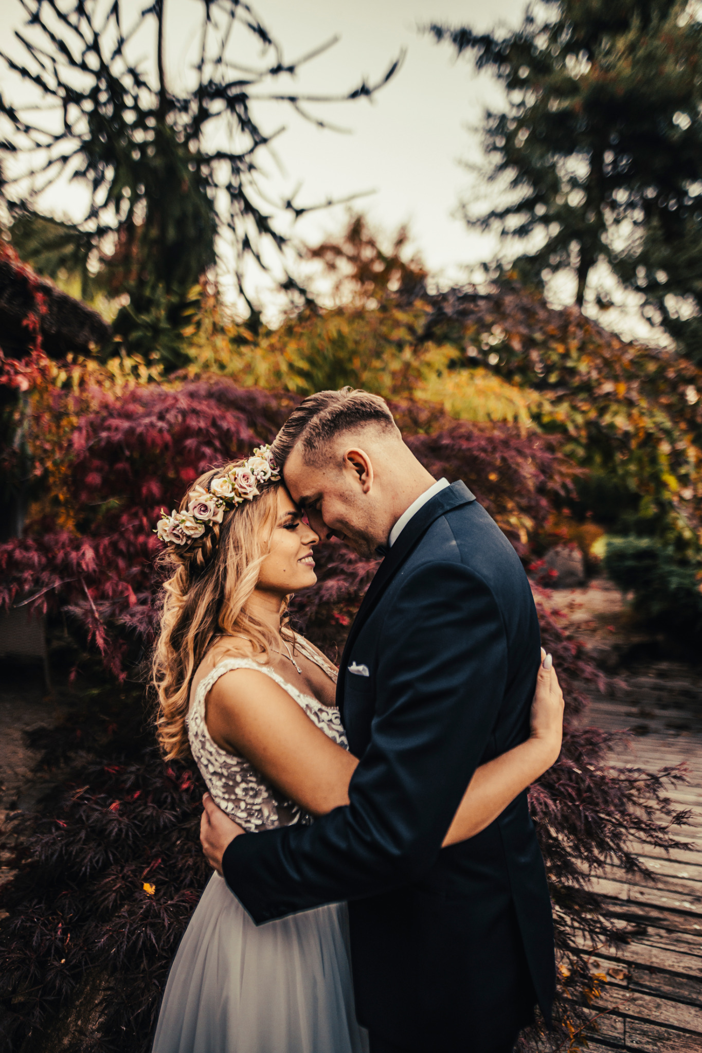 Para młoda w objęciach, panna młoda z wiankiem z róż na głowie, w tle jesienne drzewa z czerwonymi liśćmi.