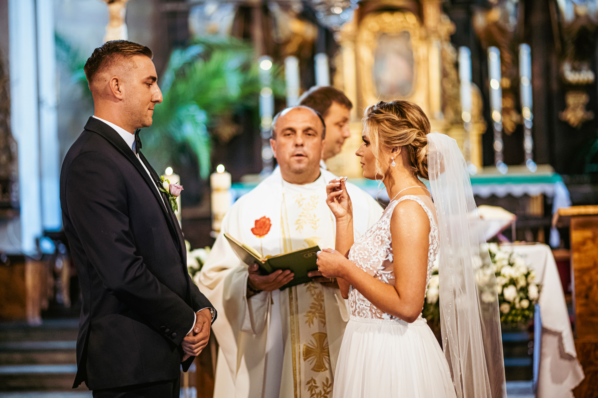 Wymiana obrączek podczas ceremonii ślubnej w kościele, panna młoda w koronkowej sukni i welonie, pan młody w ciemnym garniturze z butonierką, ksiądz z otwartą księgą.