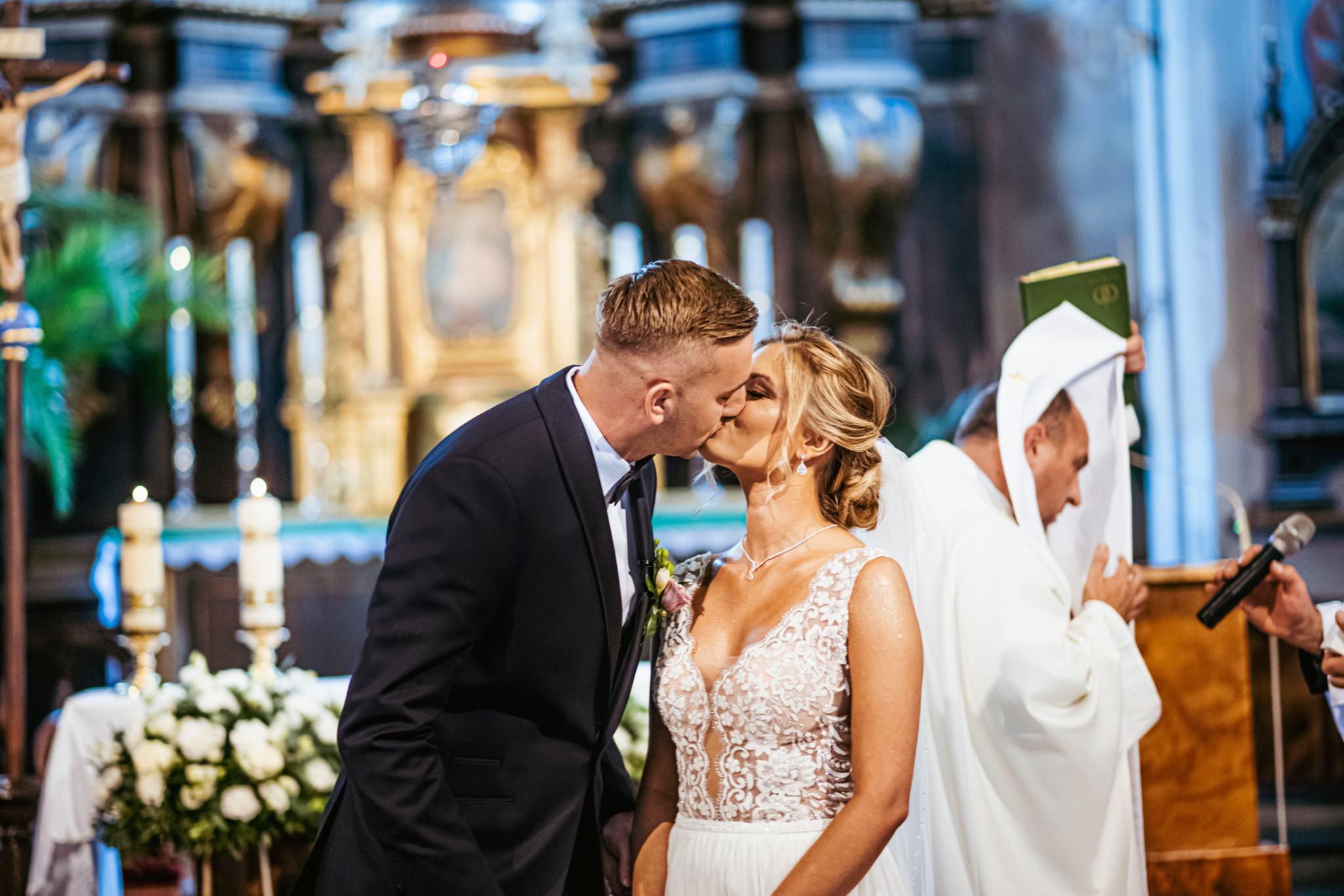 Pocałunek nowożeńców w kościele na tle ołtarza, z widocznym księdzem w białej szacie i mikrofonem.