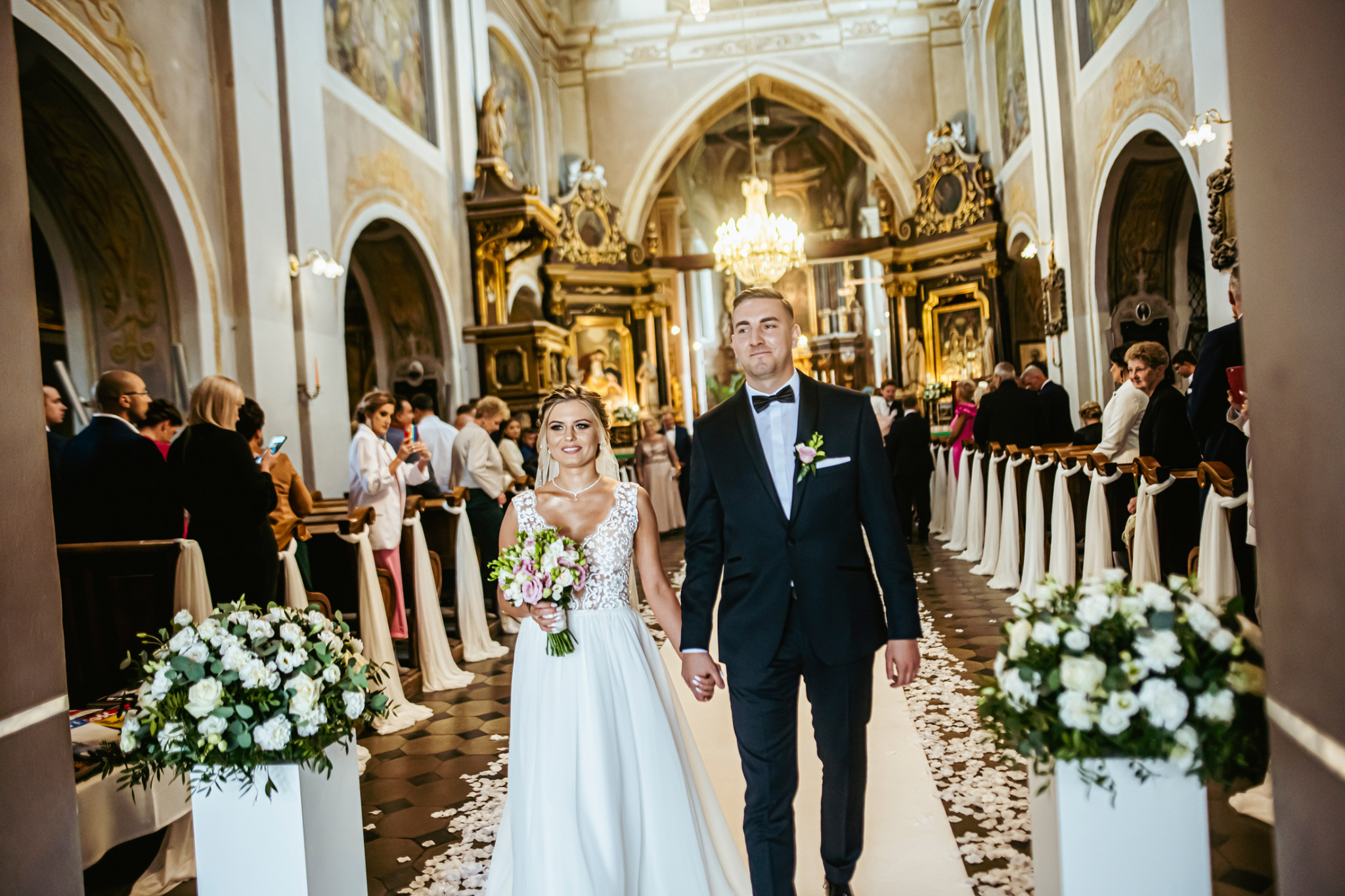 Para młoda, w sukni ślubnej z bukietem i w garniturze z muszką, wychodzi z kościoła po ceremonii, otoczona gośćmi i dekoracjami kwiatowymi.