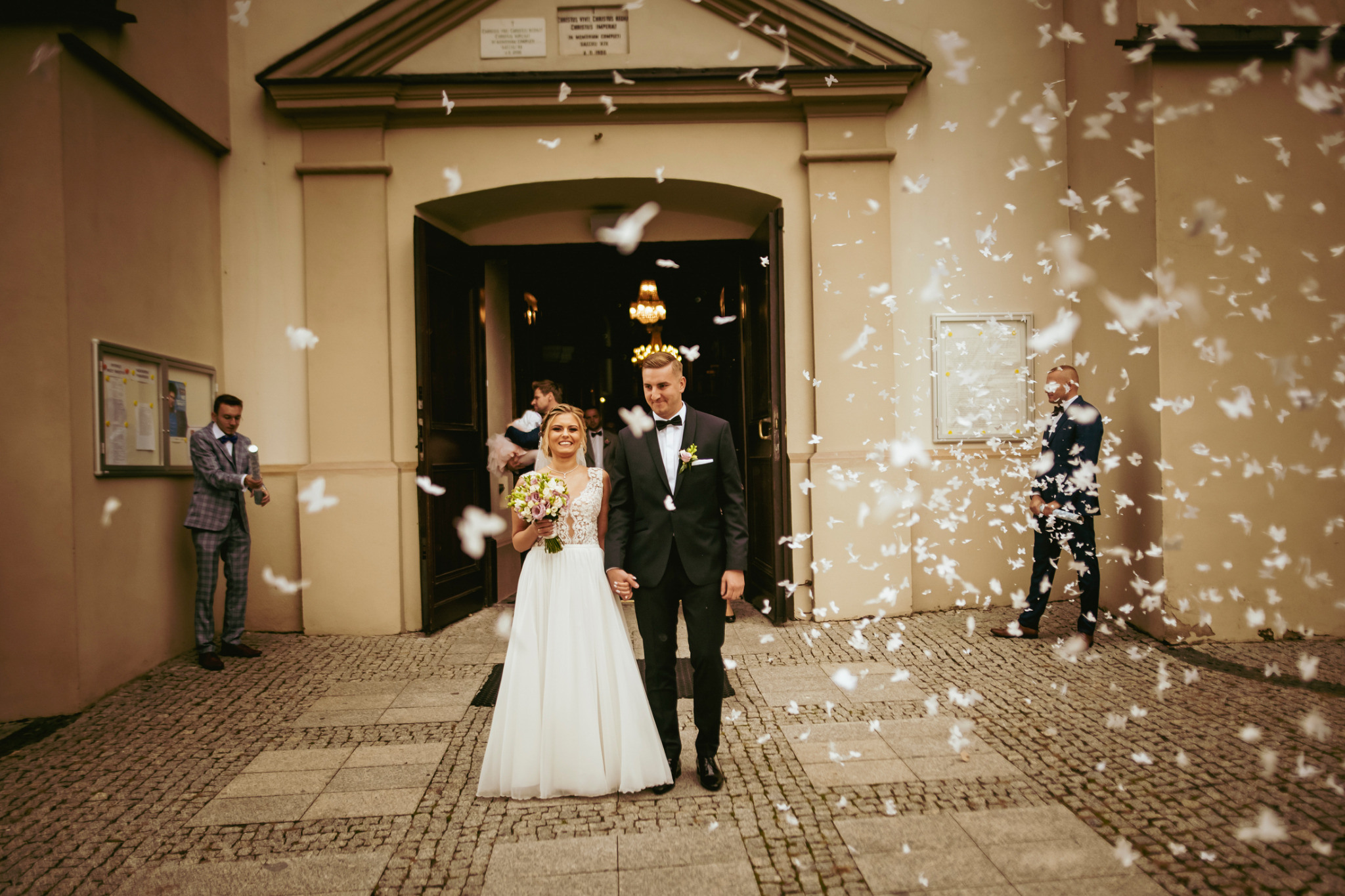Para młoda wychodzi z kościoła, obsypana białymi konfetti w kształcie motyli, w tle mężczyźni w garniturach.