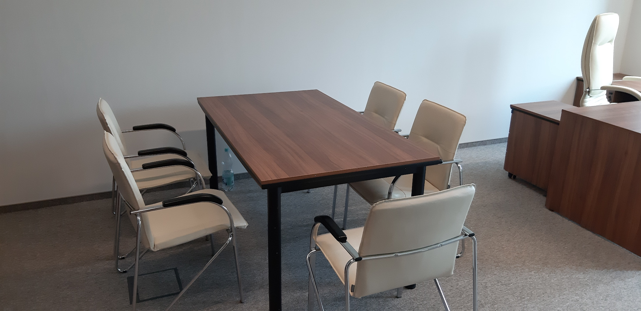 Sala konferencyjna z prostokątnym stołem z ciemnego drewna na czarnych metalowych nogach i krzesłami z jasnej skóry z czarnymi podłokietnikami, butelka wody mineralnej pod stołem.