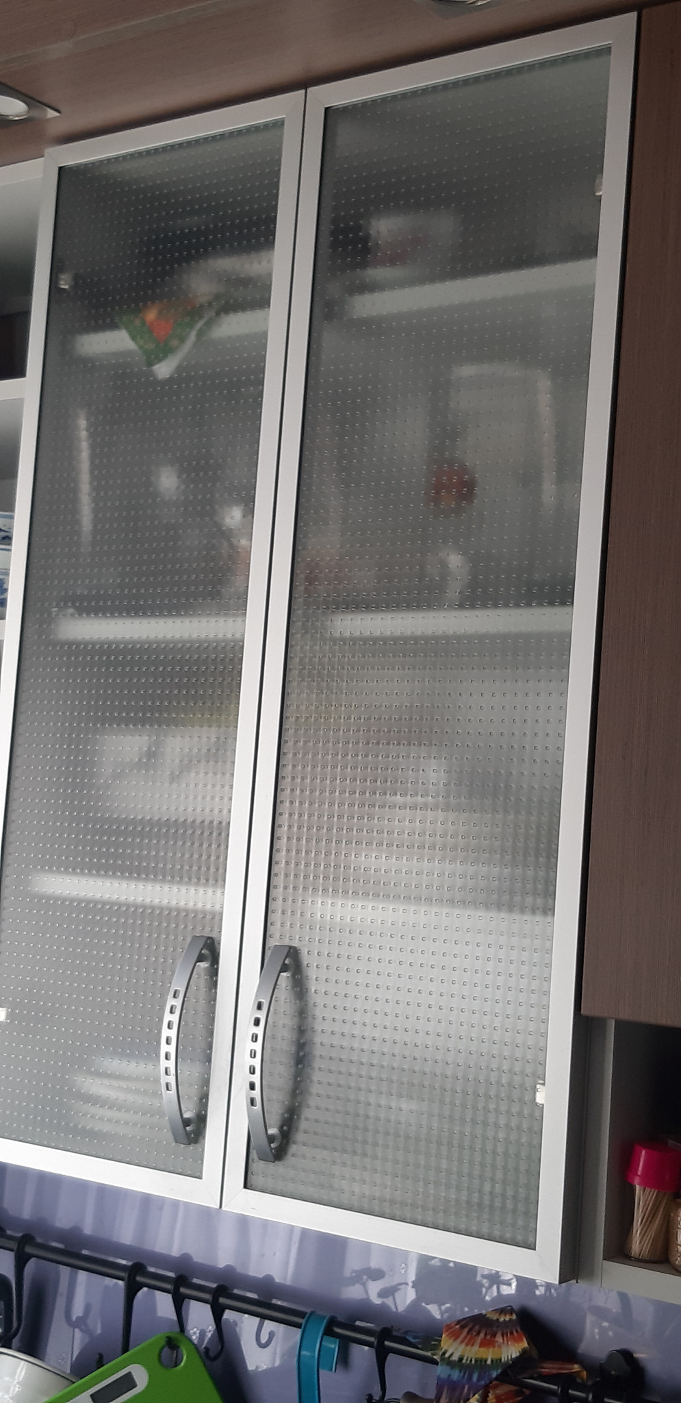 Szklana witryna w kuchni z aluminiową ramą i uchwytami, ukazująca zawartość szafki z delikatnym wzorem na szkle.